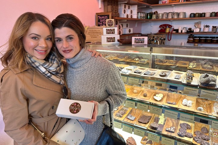 Two women smile holding chocolate boxes in front of a display case with assorted chocolates in a shop.