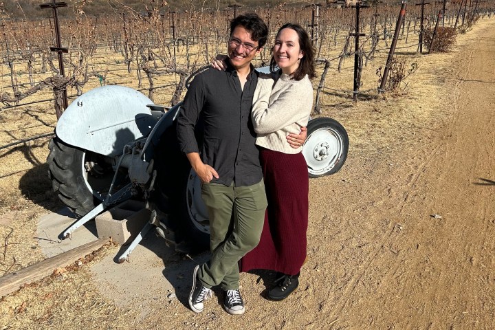 A couple poses in a vineyard, next to a small tractor, under a partly cloudy sky.