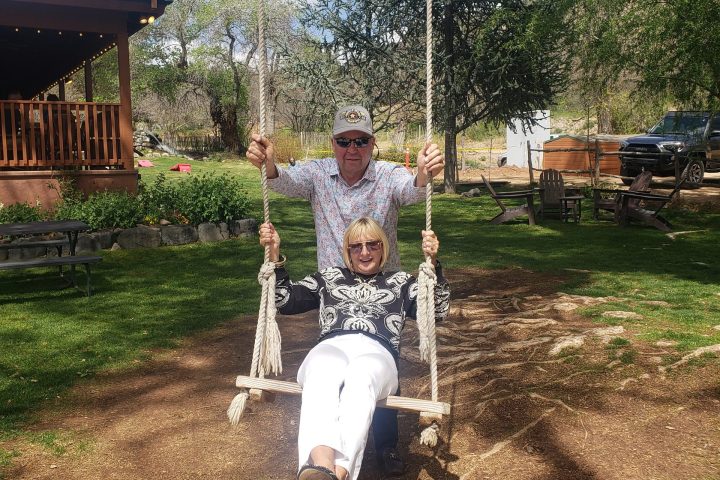 Two people on a swing outdoors with trees and a house in the background.