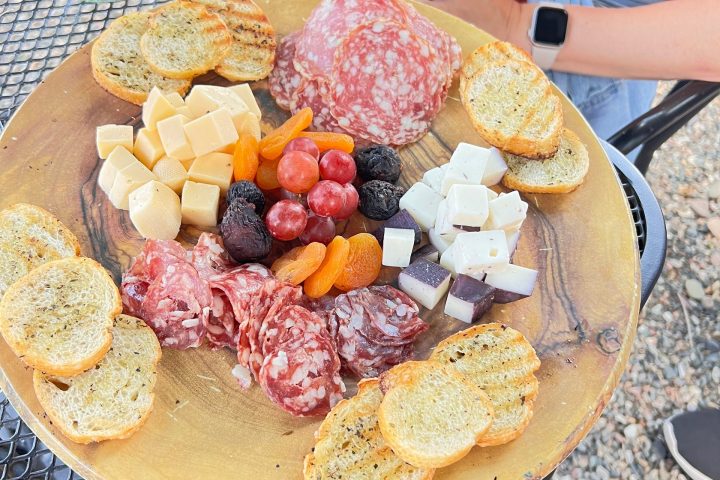 Charcuterie board with meats, cheeses, fruits, and bread slices on table with person wearing red.