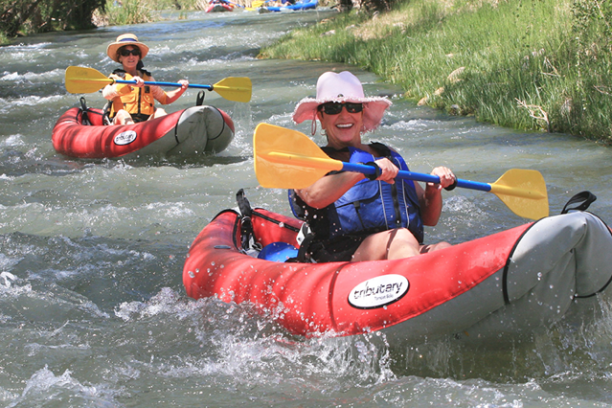 Two people kayak down a river in red inflatable kayaks, smiling and paddling with yellow and blue oars.
