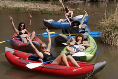 Four people in colorful canoes posing with peace signs on a calm river.