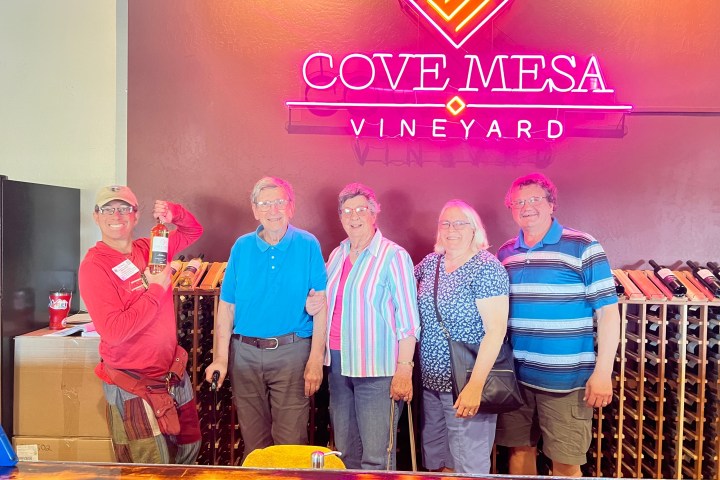 Five people smiling in front of a neon sign at Cove Mesa Vineyard with wine bottles displayed.