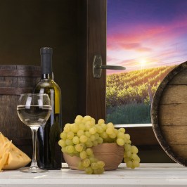 Wine bottle, glass, cheese, grapes on table with barrel; vineyard sunset view through window.