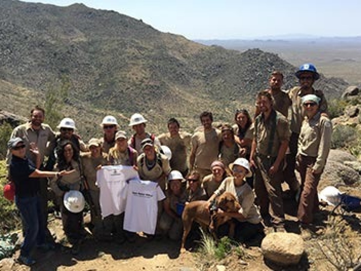 Group of people and a dog posing on a mountain trail holding a T-shirt with a scenic view in the background.