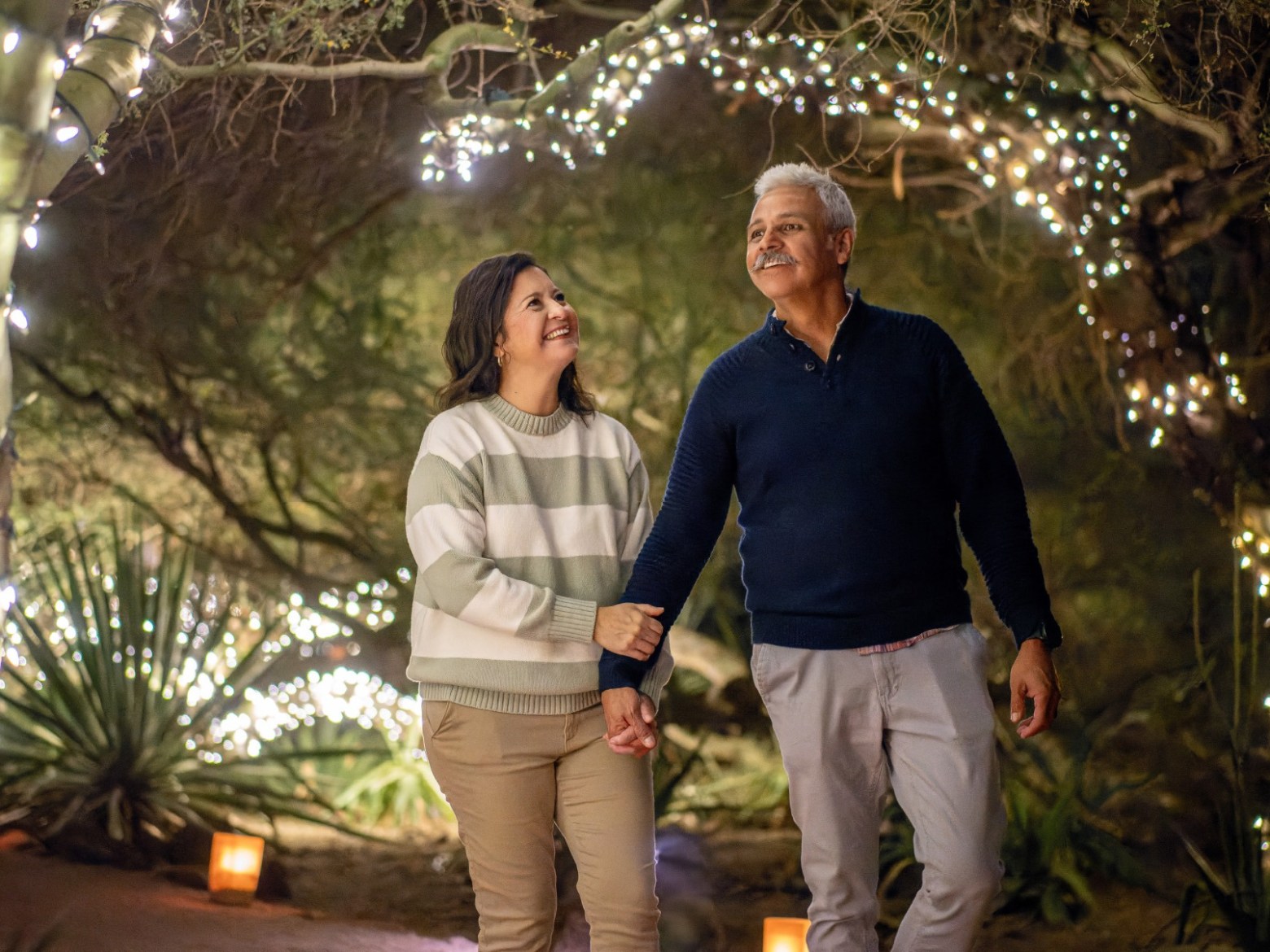Couple walking under trees adorned with string lights at night.