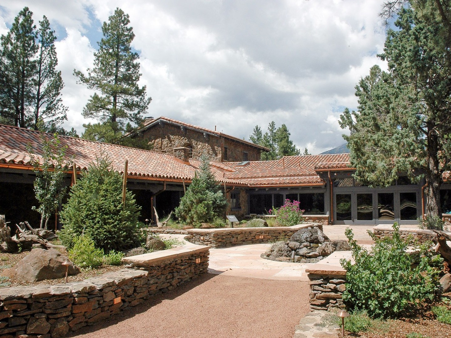 Stone building with terracotta roof surrounded by trees under a cloudy sky.