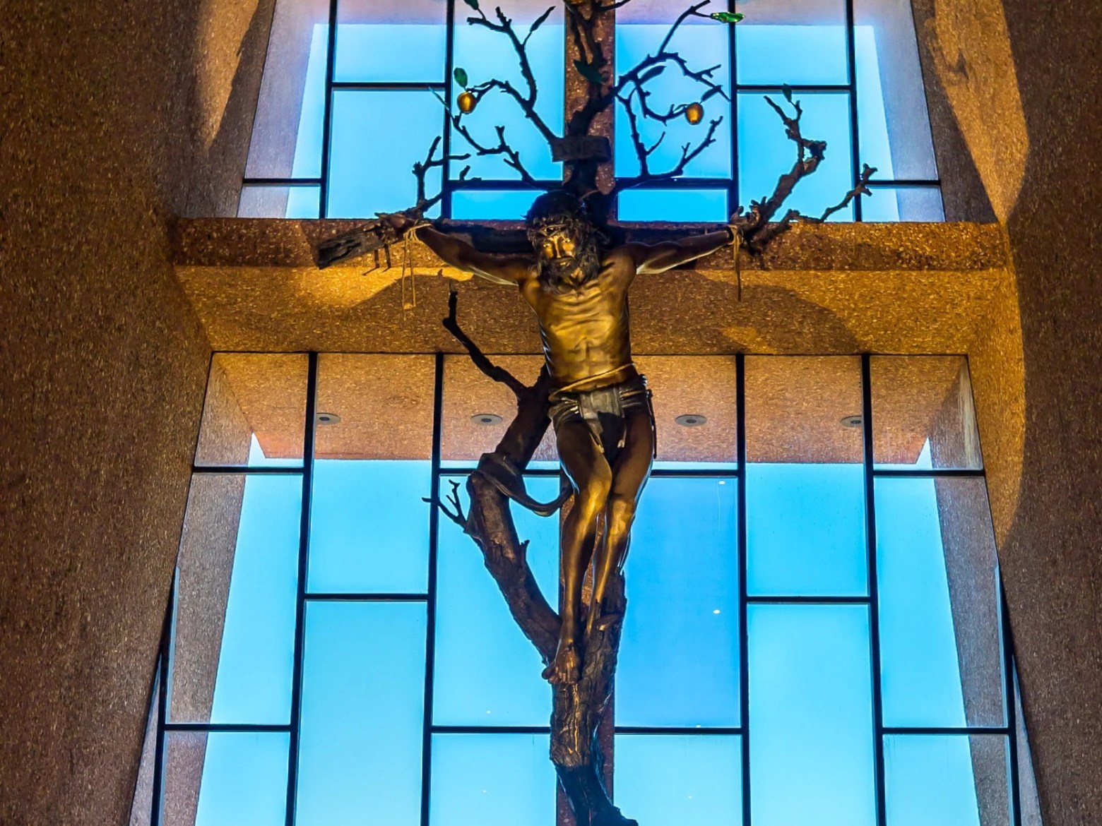 Crucifix sculpture in church with a large window and blue sky backdrop.