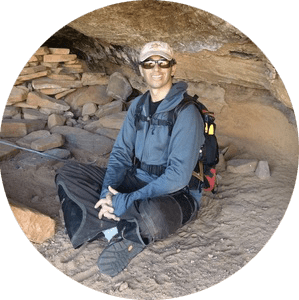 Person sitting in cave with backpack, wearing sunglasses and cap.