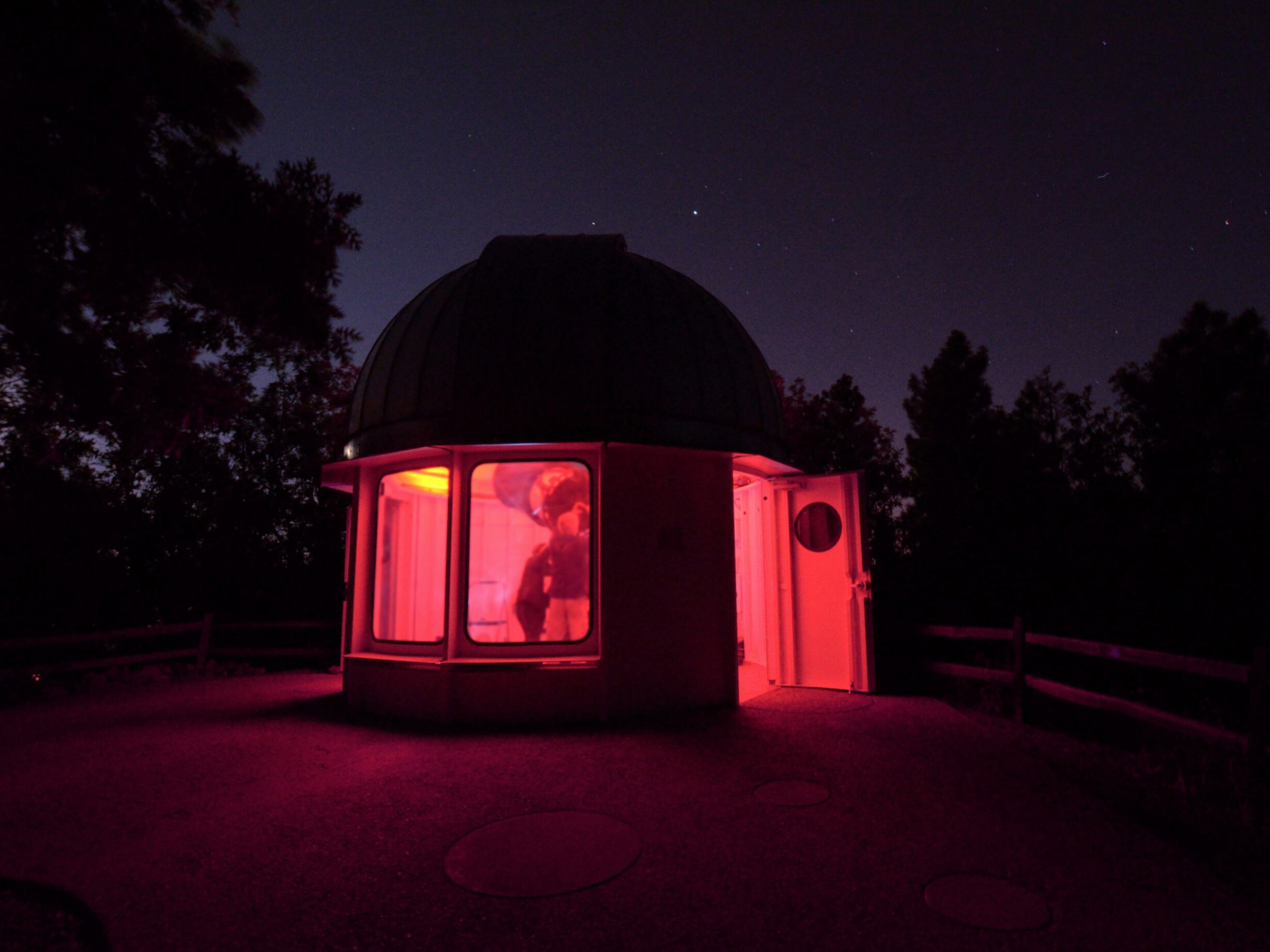 Small observatory at night with red-lit interior and open door, surrounded by trees.
