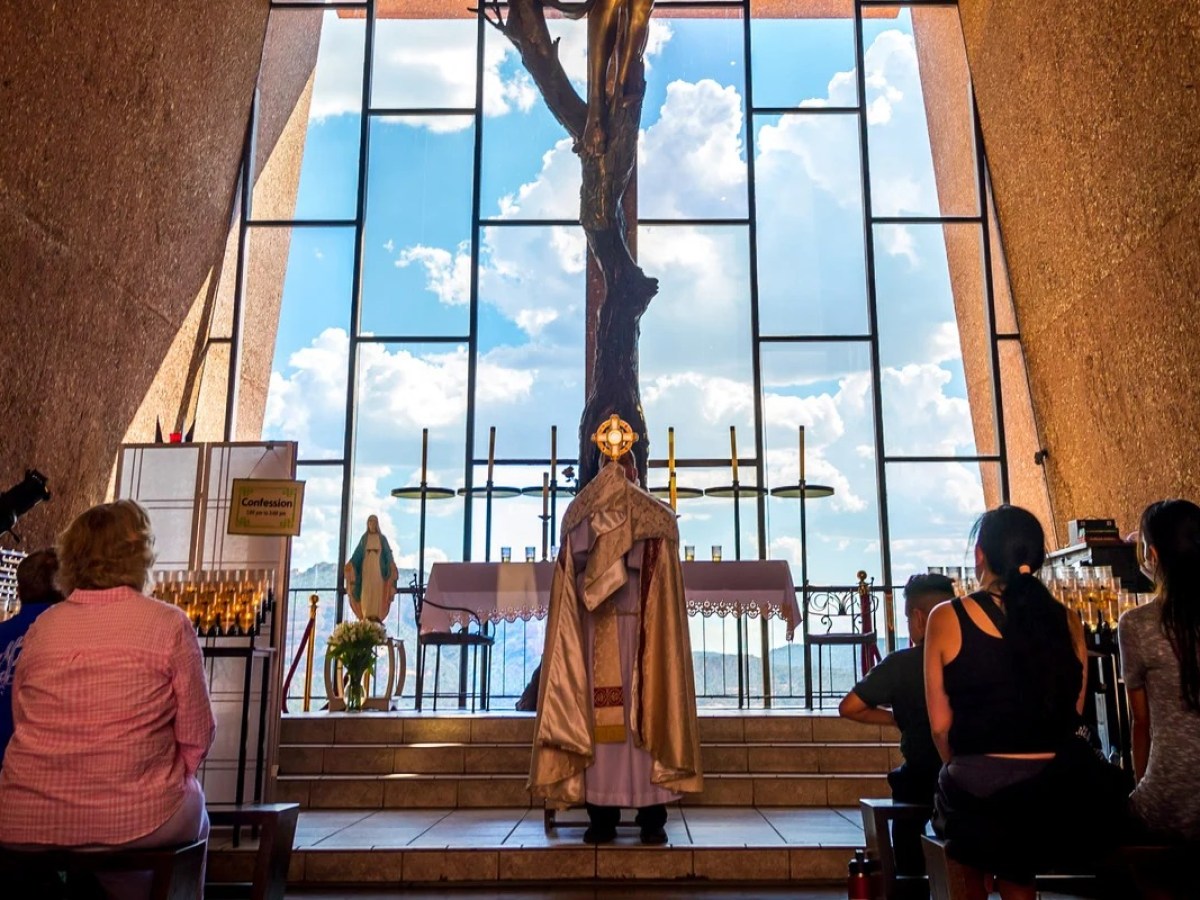Priest leads service in church with stained glass window and seated congregation.