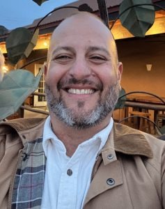 Smiling man in jacket outdoors with leaves and building in background.
