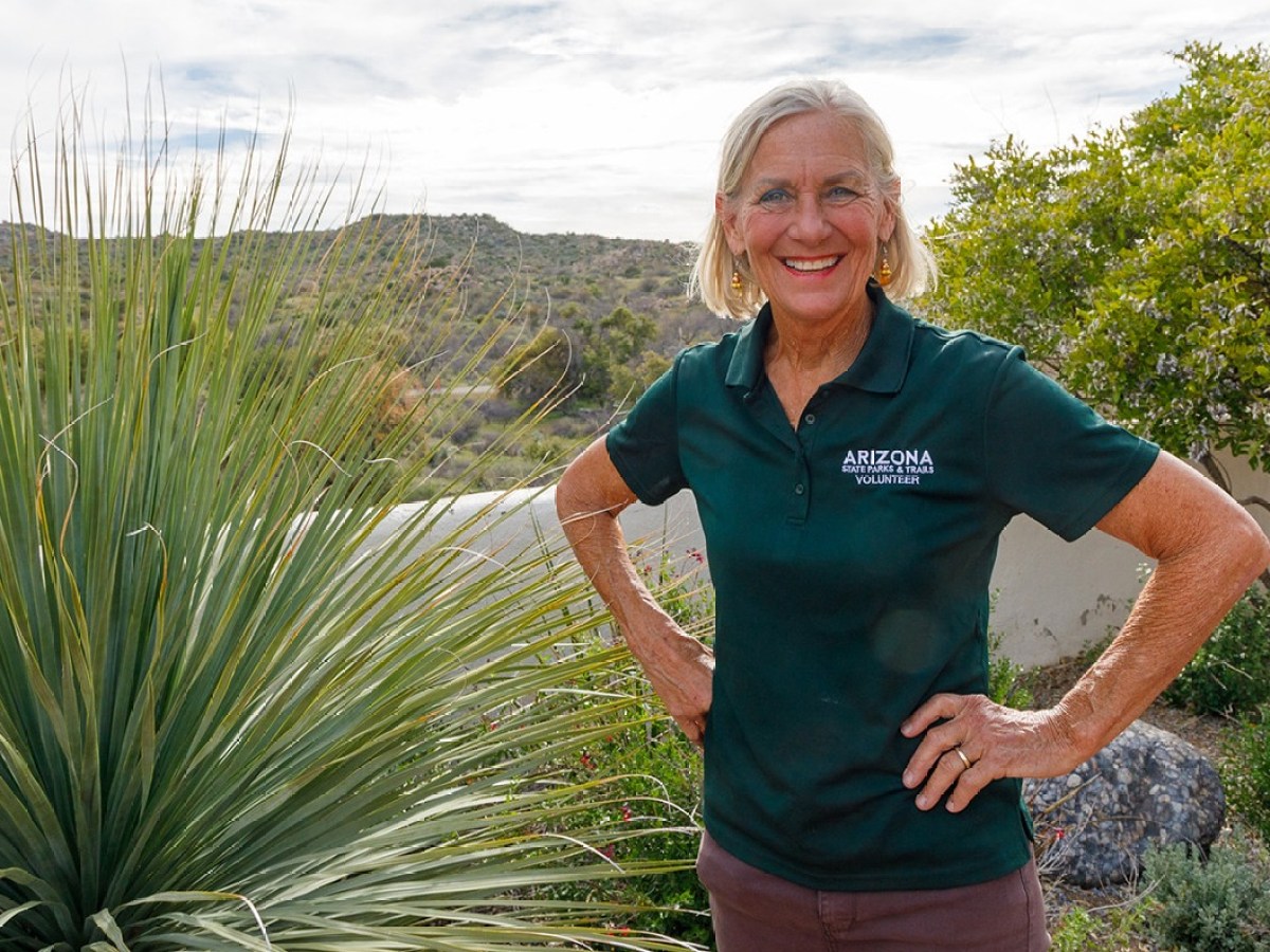 Smiling woman in a green volunteer polo shirt stands outside near desert plants.