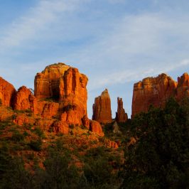 Red rock formations under a blue sky with scattered clouds, surrounded by green vegetation.