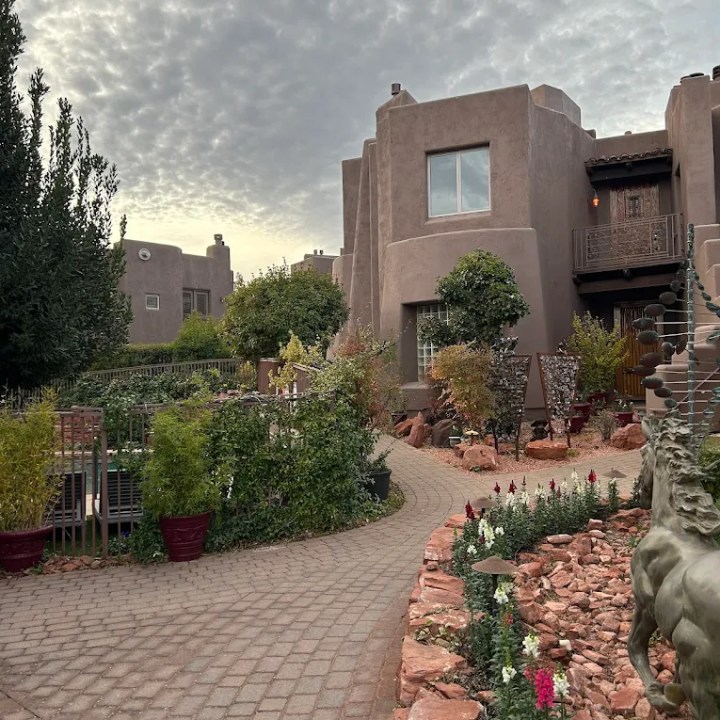 Pathway through garden leading to adobe-style buildings under cloudy sky.