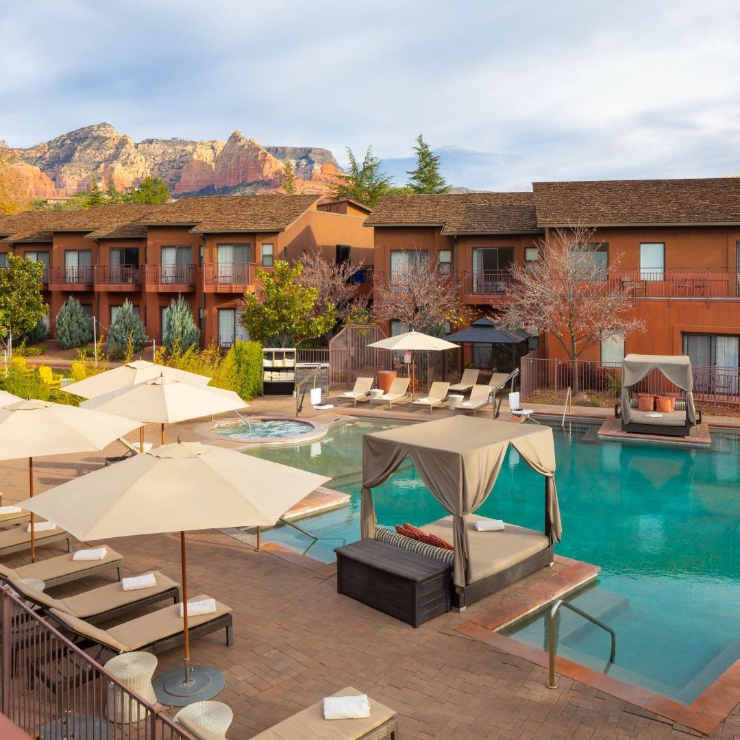 Resort pool area with loungers and umbrellas, surrounded by buildings and mountains.
