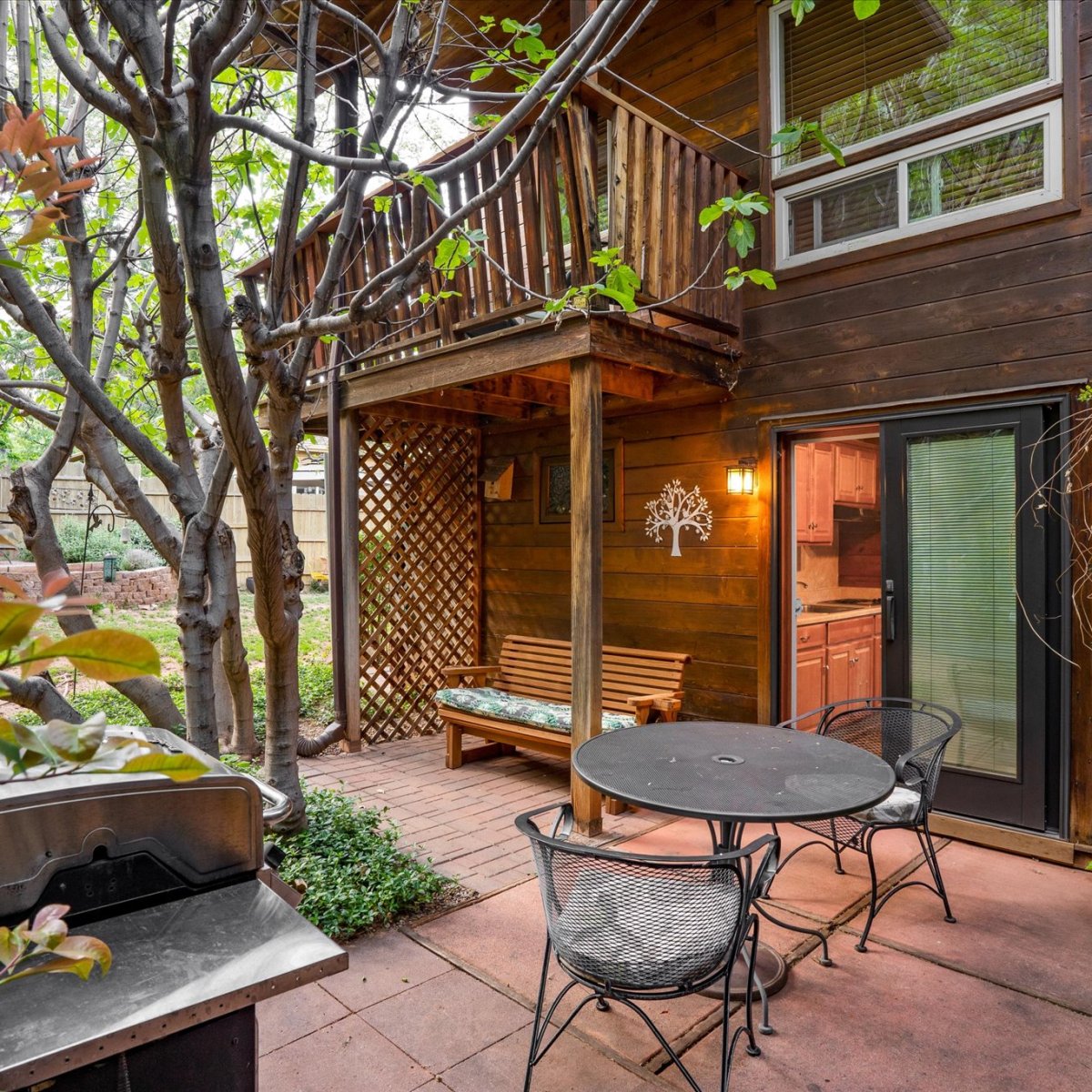 Cozy patio with grill, round table, chairs, wooden deck above, surrounded by trees and bushes.