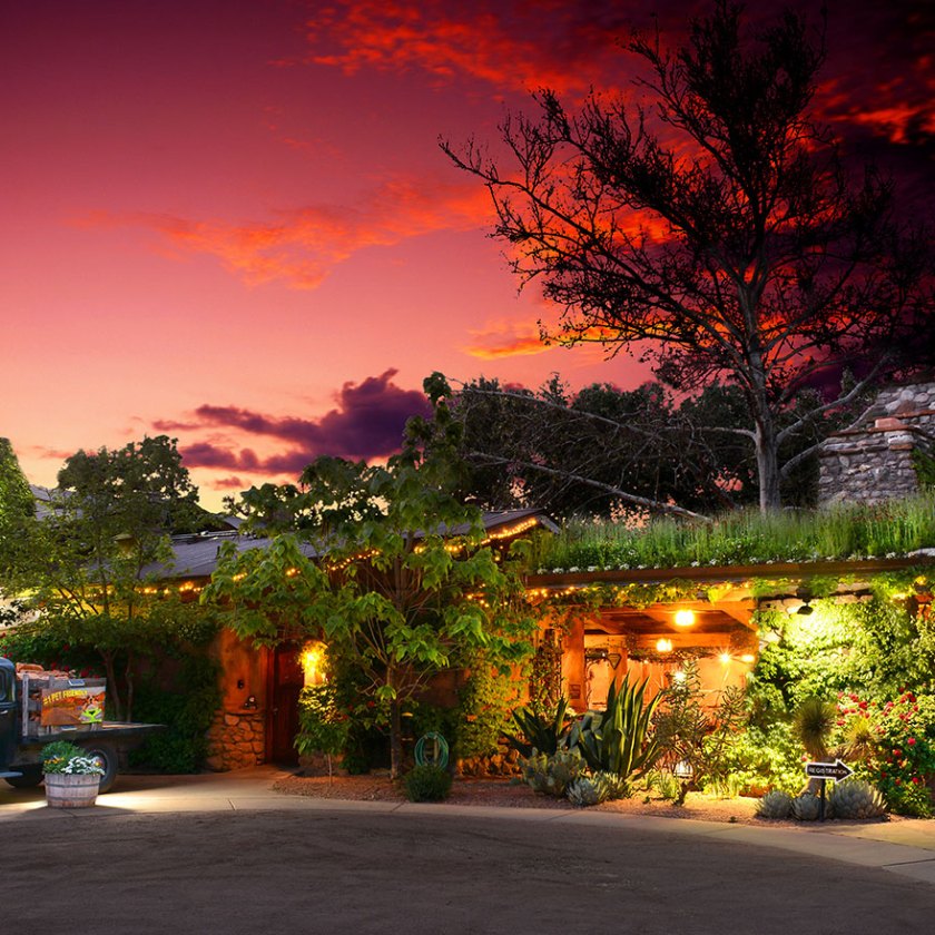 Old truck parked by rustic house under vibrant sunset sky with green foliage.