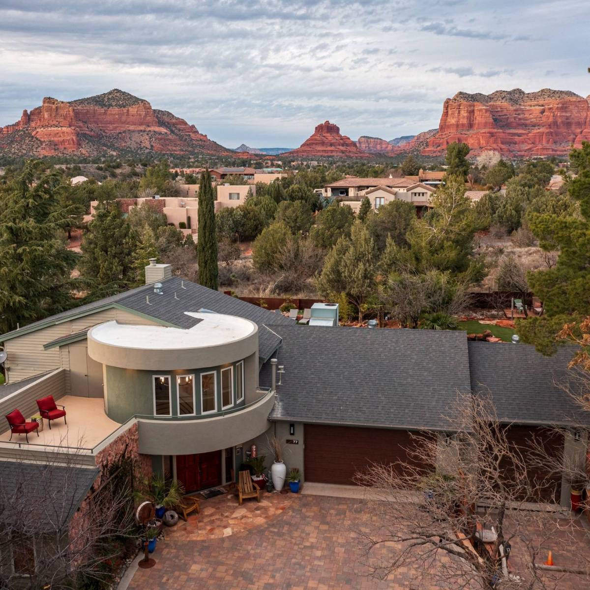 Modern house with circular balcony, surrounded by trees, with red rock formations in the background.