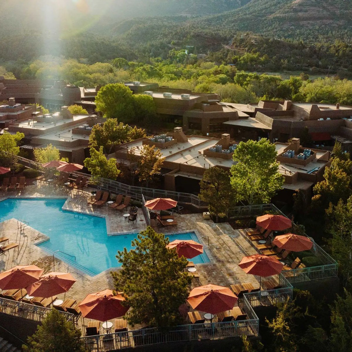 Aerial view of resort with large pool and red umbrellas, surrounded by trees and mountains.