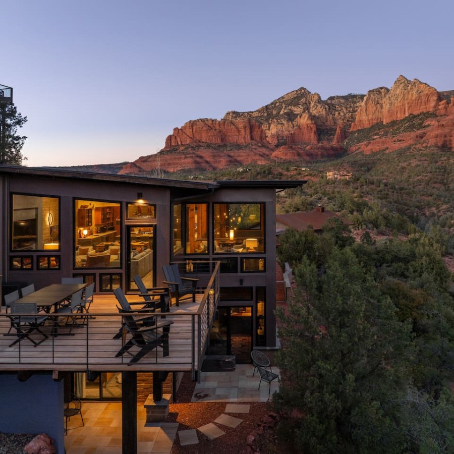 Modern house with large deck, nestled in a forested area with red rock mountains in the background at dusk.