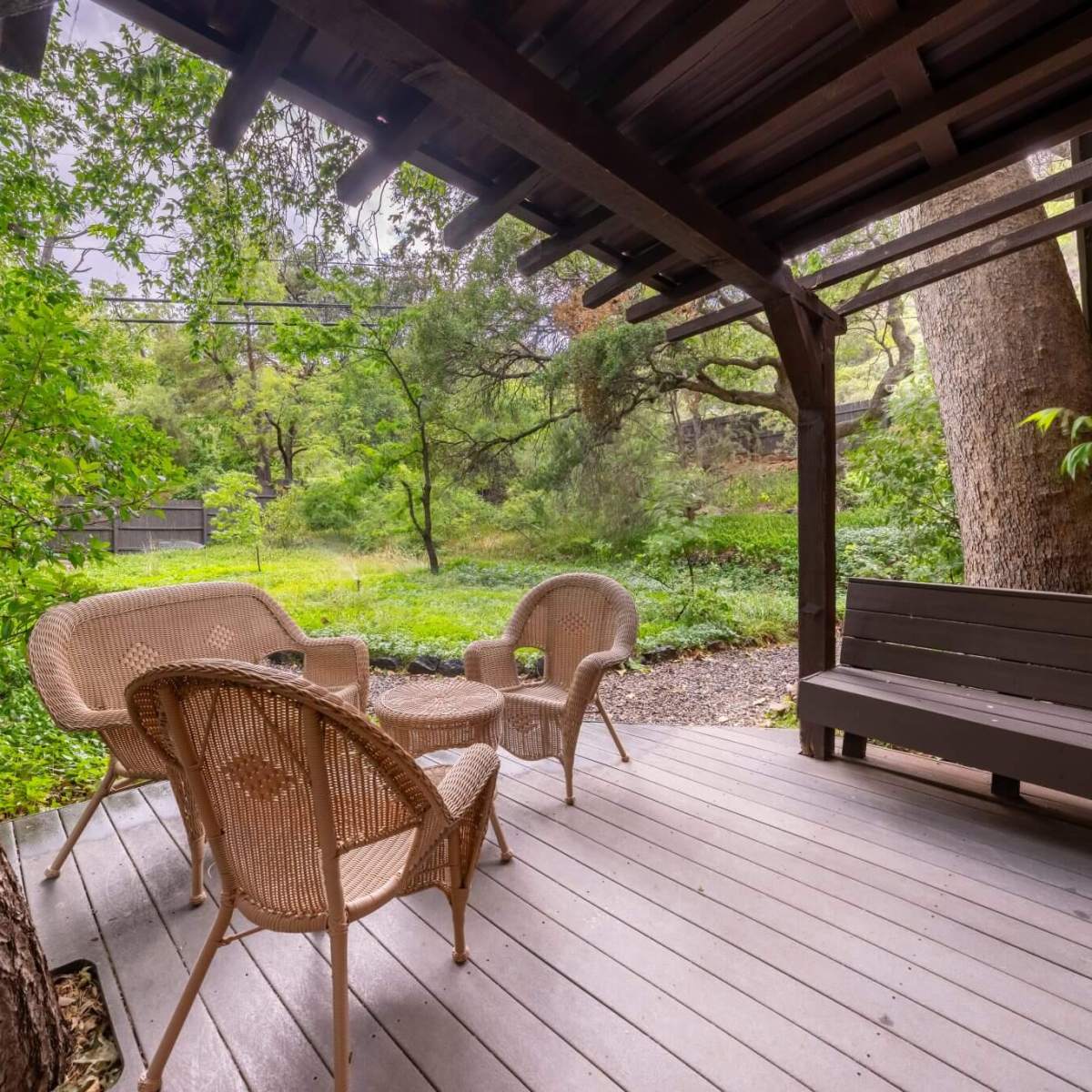 Covered wooden deck with wicker chairs and bench, surrounded by lush greenery.