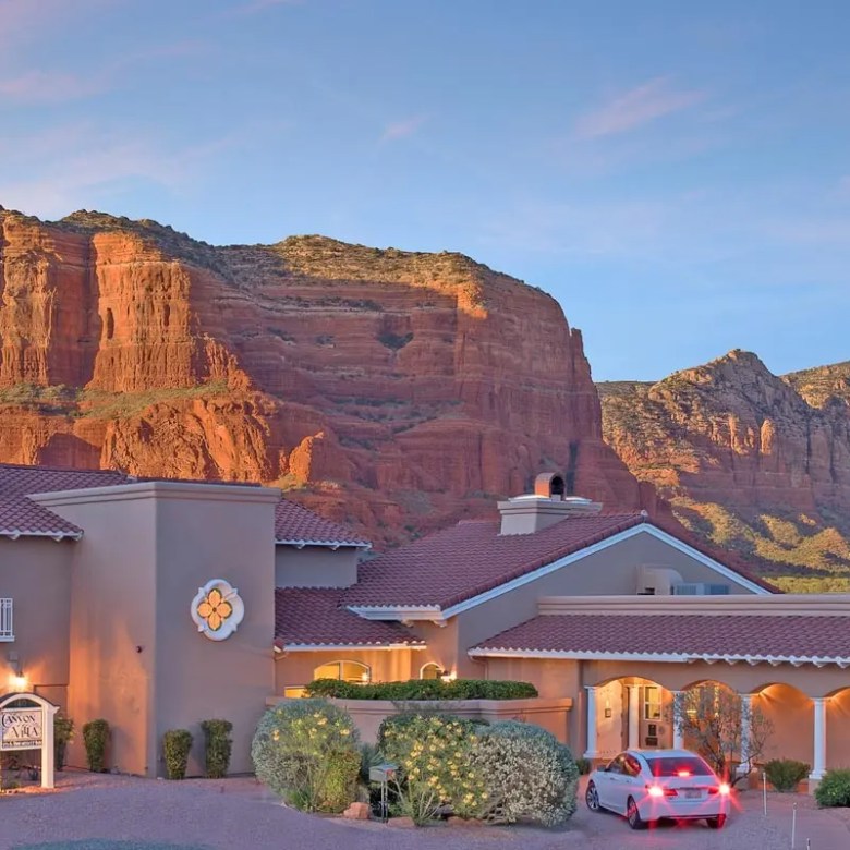 House with red rock mountains in the background and a car parked in front, under a clear sky at sunset.
