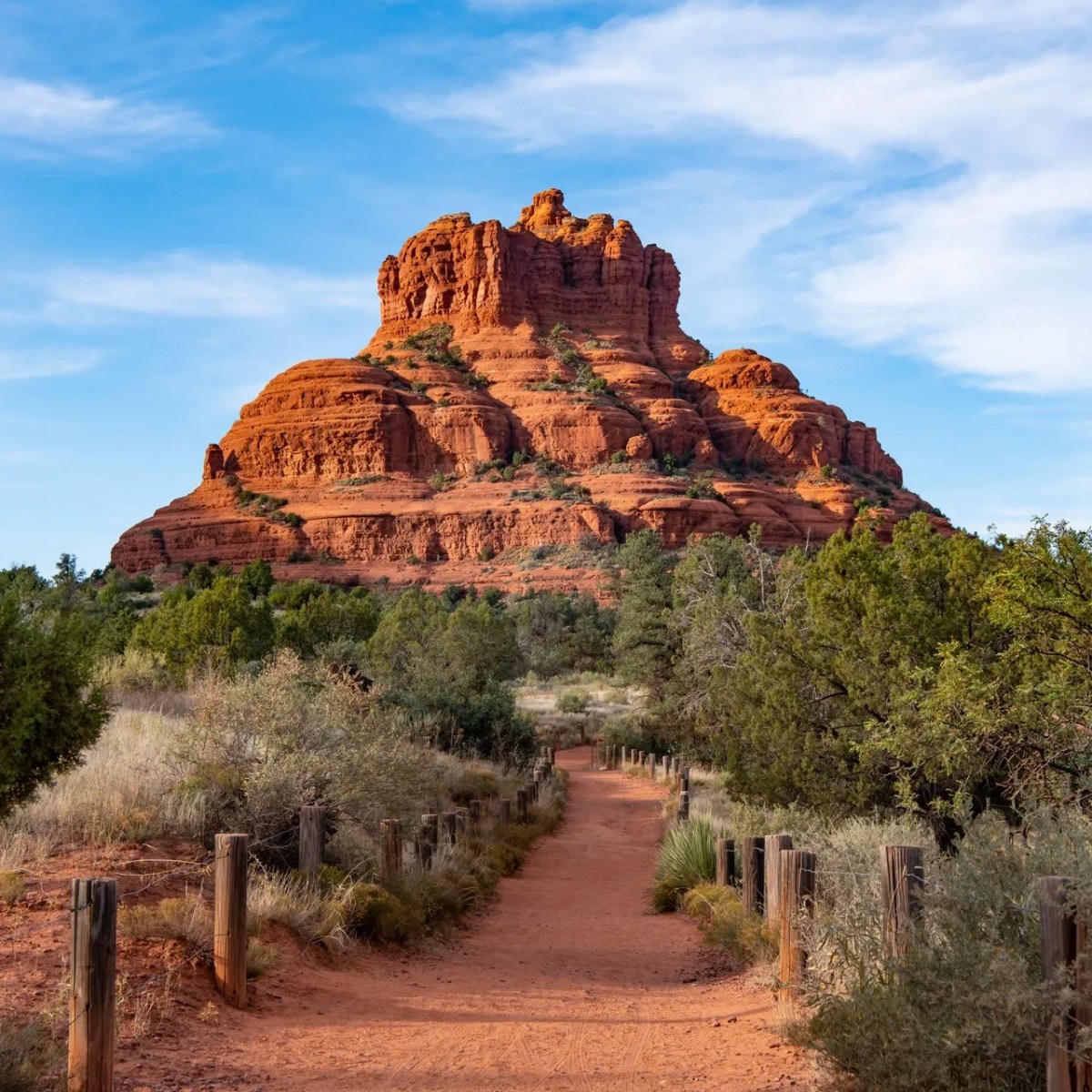 Red rock formation under blue sky, surrounded by trees and a dirt path leading towards it.