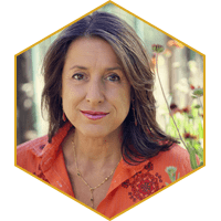 Woman with brown hair in orange shirt, smiling in garden setting, inside a hexagon frame.
