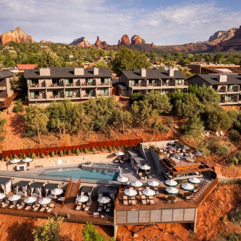 Aerial view of a resort with a pool, surrounded by trees and red rock mountains under a partly cloudy sky.