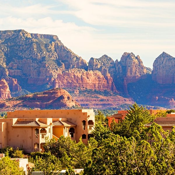 Adobe-style buildings amid trees with red rock mountains in background.